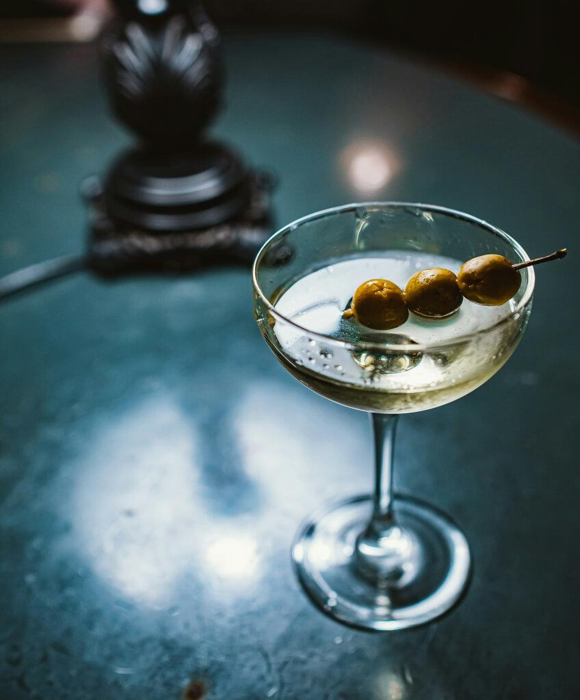 An above shot of a light yellow drink with toothpick of three olives perched on top.