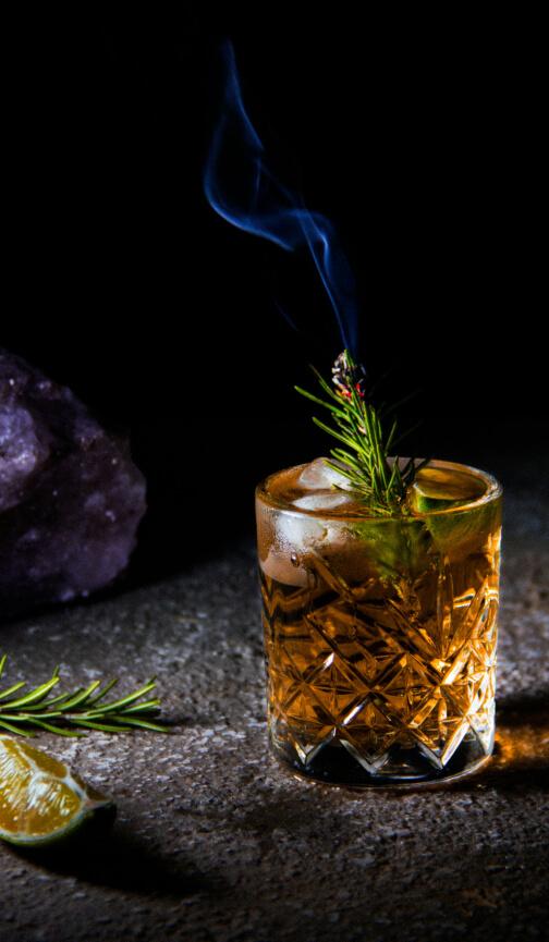 A front shot of a glass of whisky with a smoking rosemary sprig and ice inside it. Next to the glass is a slice of lime and a rosemary sprig.