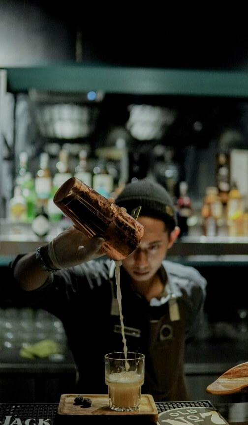 A focused shot of a bartender pouring a drink from a mixer. The camera focus is on the mixer and pouring liquid, with the bartender and background bar blurred.